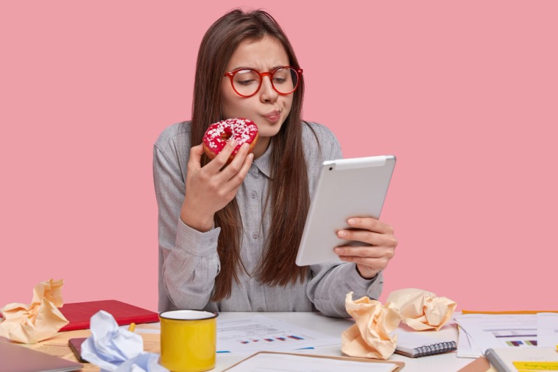 chica seria sostiene deliciosa rosquilla mientras trabaja con tablet